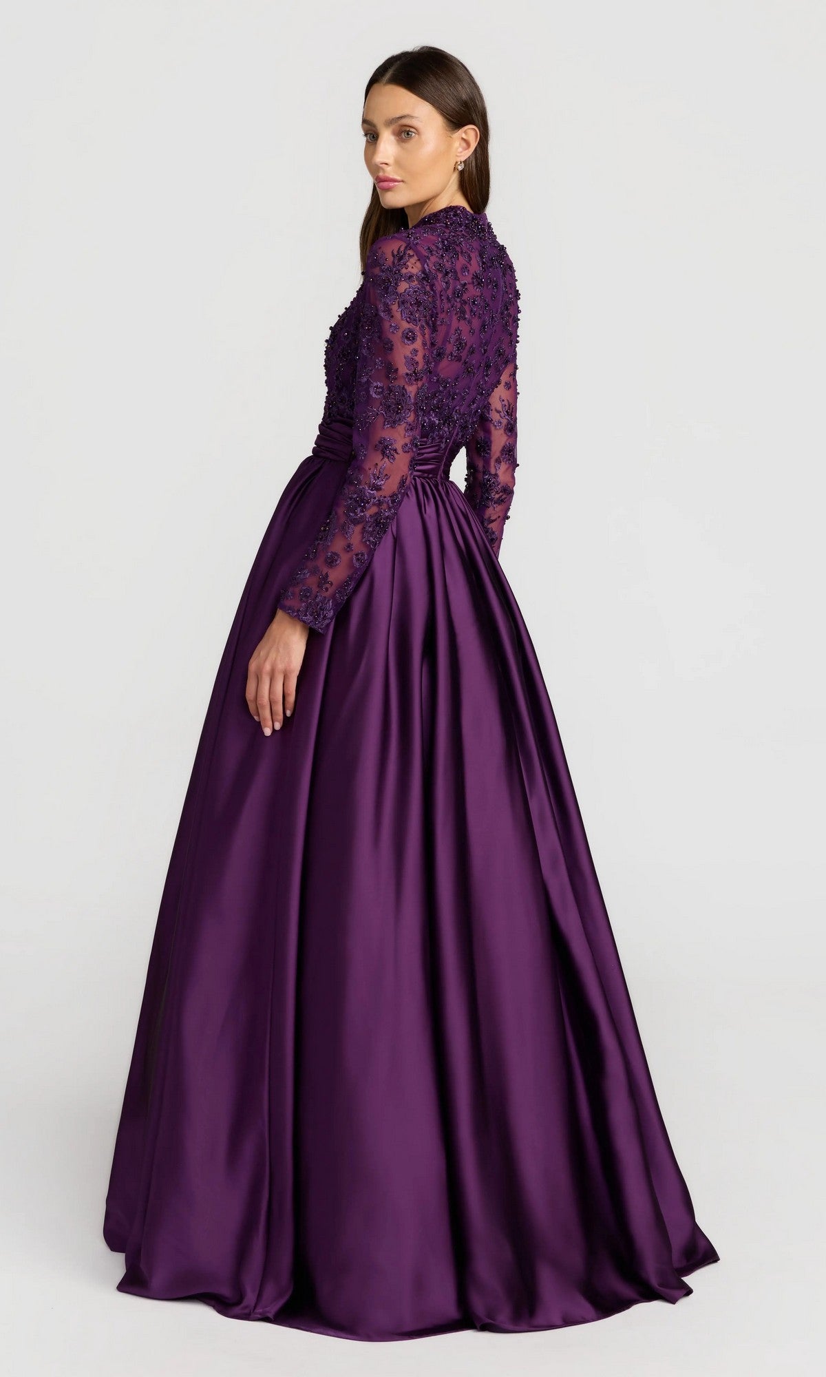 A woman in the Mac Duggal 21189 long formal dress, featuring long sleeves, a beaded-embroidered bodice, and a satin floor-length skirt, stands slightly turned from the camera against a plain background.
